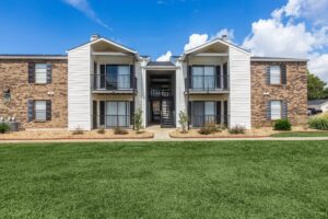 Exterior view of the two-story Edgewater Pointe Apartments building in Biloxi, MS, featuring a mix of brown brick and light siding, private balconies with black railings, and a manicured green lawn under a blue sky.