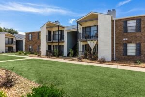 Exterior view of two-story apartment buildings at Edgewater Pointe in Biloxi, MS, featuring a mix of brick and beige siding, private balconies, patios, and a manicured green lawn under a blue sky.