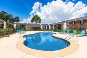Kidney-shaped swimming pool with turquoise lounge chairs on the sun deck, surrounded by a black fence and brick apartment buildings at Edgewater Pointe Apartments in Biloxi, MS.