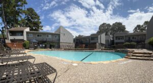 Kidney-shaped community swimming pool with natural rock edging and multiple brown lounge chairs arranged around the pebble deck at Trailwood Village Apartments complex on a sunny day.