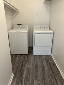In-unit laundry room at Trailwood Village Apartments in Kingwood, TX, featuring a full-size white top-load washer and front-load dryer on wood-style flooring.