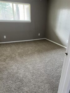 Spacious empty bedroom in a 3-bedroom Abberly floor plan at Trailwood Village Apartments in Kingwood, TX, featuring grey carpeting and a large window.